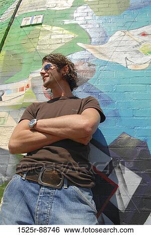 Low angle view of a young man standing with his arms crossed in front of a graffiti covered wall View Large Photo Image Stock Photograph - Low angle view of a young man standing with his arms crossed in front of a graffiti covered wall. Fotosearch
