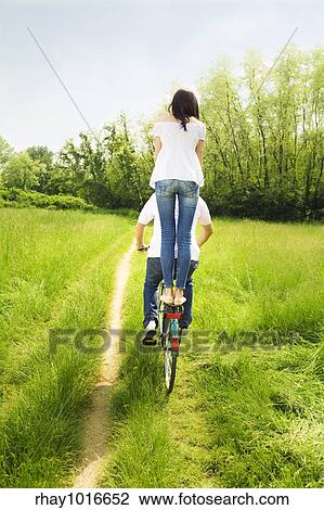 Stock Image - couple biking in park. Girl standing on the back of the bike. Fotosearch