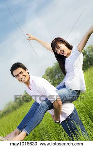 Stock Image - "Man giving woman piggyback in meadow, laughing. Narrow focus on his eye. ". Fotosearch