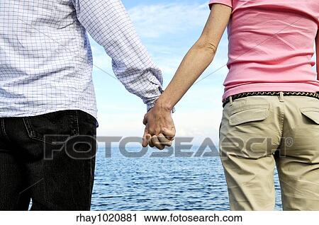 Stock Image - Mature romantic couple holding hands on the beach. Fotosearch