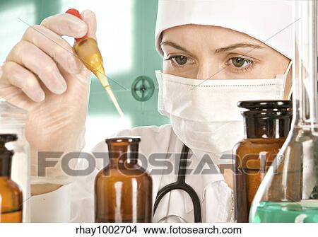 Picture - The woman-doctor with concentration drips from a pipette in a small bottle near a table on which there is a flask with a greenish liquid The woman-doctor with concentration drips from a pipette i.... Fotosearch