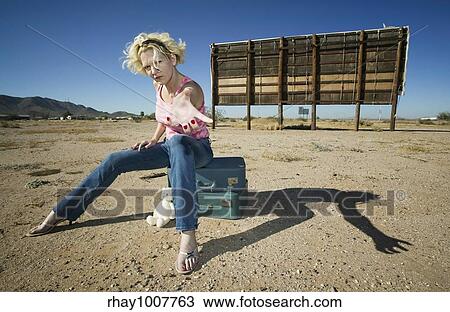 Stock Image - Woman sitting on suitcases in front of an old billboard reaching to the camera. Fotosearch