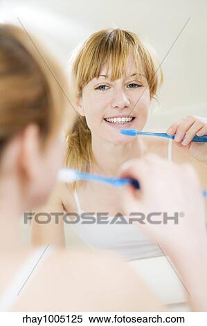 Stock Photography - Young woman looking into a mirror brushing her teeth. Fotosearch