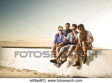 Stock Image - Friends sitting on wall with smartphone, Mission Beach, San Diego, California, USA. Fotosearch