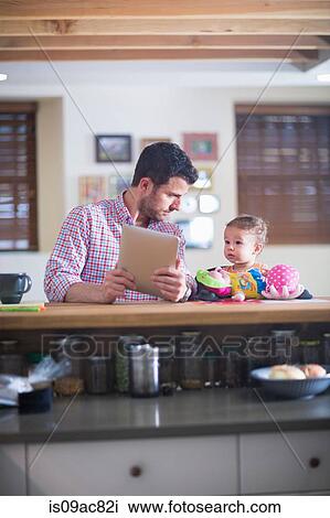 Man and baby sitting at kitchen counter looking at digital tablet View Large Photo Image Stock Image - Man and baby sitting at kitchen counter looking at digital tablet. Fotosearch