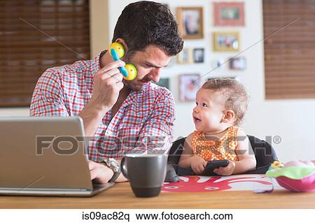 Stock Photography - Man and baby sitting at kitchen counter playing with smartphone and toy phone. Fotosearch