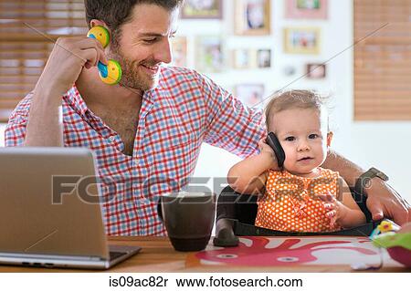 Stock Photography - Man and baby sitting at kitchen counter playing with smartphone and toy phone. Fotosearch