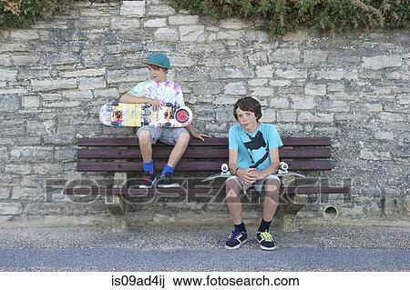 Stock Image - Portrait of two boys sitting on bench holding skateboards. Fotosearch