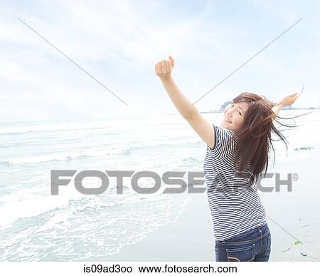 Picture - Portrait of young woman on beach with arms raised, looking over shoulder. Fotosearch