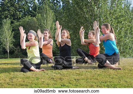 Stock Image - Teenage girls and tutor sitting on grass doing yoga. Fotosearch
