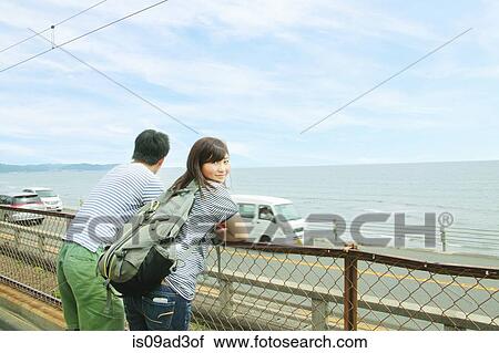 Stock Image - Young couple leaning against fence, looking out to sea. Fotosearch