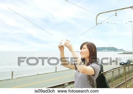 Stock Image - Young woman taking photo at coast with smartphone. Fotosearch