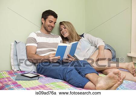 Stock Image - Couple relaxing on bed looking at book. Fotosearch