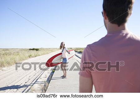 Stock Photography - Couple with surfboard on coastal path, Breezy Point, Queens, New York, USA. Fotosearch