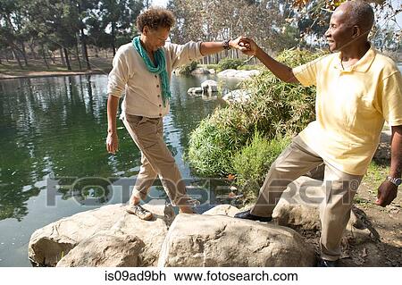 Stock Image - Portrait of senior couple stepping on rocks by lake in park. Fotosearch