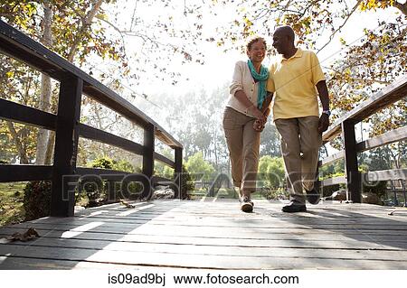 Stock Image - Senior couple walking on footbridge in park. Fotosearch