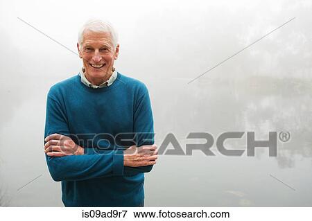 Stock Photo - Senior man with arms crossed by foggy lake. Fotosearch