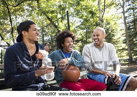 Stock Image - Young men sitting on park bench laughing. Fotosearch