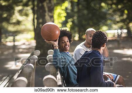 Stock Image - Young men sitting on park, one holding basketball. Fotosearch