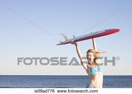 Stock Photograph - Young woman holding surfboard up, Breezy Point, Queens, New York, USA. Fotosearch