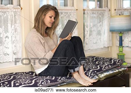 Stock Image - Young woman reading book in window seat. Fotosearch