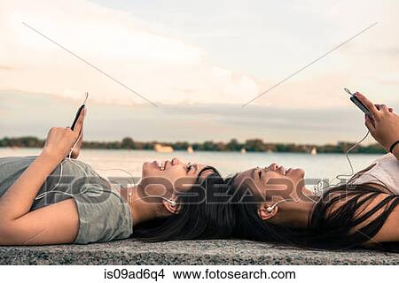 Young women lying head to head at waterfront listening to music through earphones View Large Photo Image Picture - Young women lying head to head at waterfront listening to music through earphones. Fotosearch