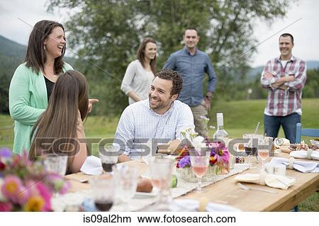 Picture - Family and friends gathered around, talking, at the end of a meal, outdoors. Fotosearch