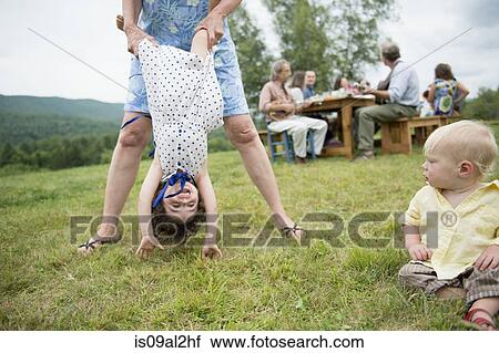 Stock Image - Female family member playfully holding toddler by legs at family gathering, outdoors. Fotosearch