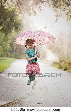 Stock Image - Girl holding up umbrella and jumping puddles on street. Fotosearch