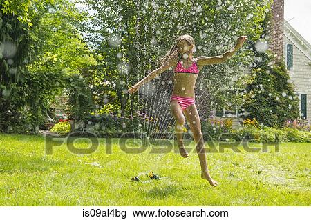 Stock Image - Girl in swimming costume jumping over garden sprinkler. Fotosearch