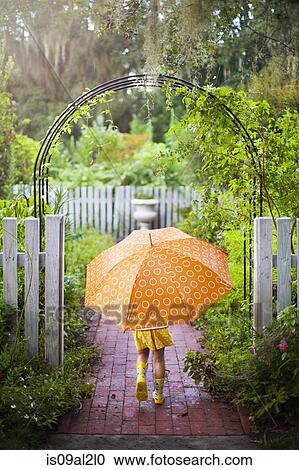Stock Image - Girl walking through garden gate carrying umbrella. Fotosearch