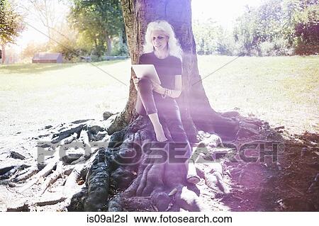 Stock Image - Gray haired woman taking break reading digital tablet in city park. Fotosearch