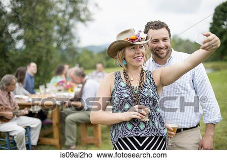 Picture - Man and woman taking self portrait with smartphone while family friends sit at table behind, outdoors. Fotosearch