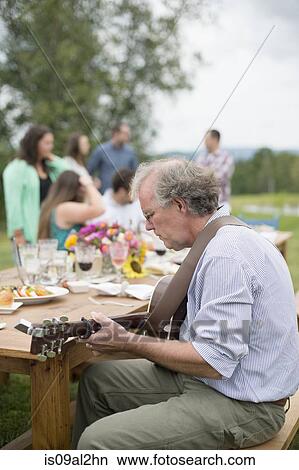 Mature man playing guitar while friends and family talk together after meal, outdoors View Large Photo Image Picture - Mature man playing guitar while friends and family talk together after meal, outdoors. Fotosearch