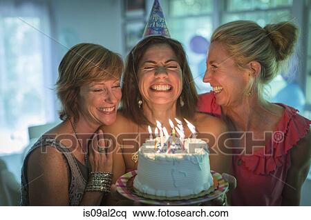 Stock Photography - Mature woman holding birthday cake, making wish while two friends look on. Fotosearch