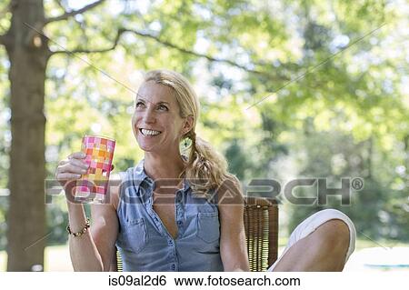 Stock Photograph - Mature woman sitting in garden with drink. Fotosearch