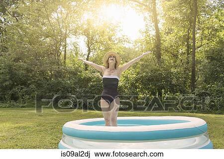 Stock Image - Mature woman standing in paddling pool, arms outstretched. Fotosearch