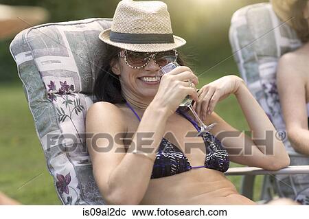 Stock Image - Mature woman wearing bikini, relaxing on sun lounger, drinking wine. Fotosearch