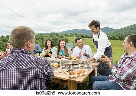 Stock Photography - Mid adult man in apron, serving plate of food to family members at table, outdoors. Fotosearch