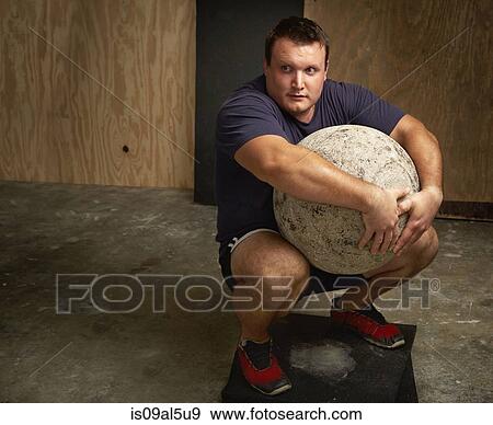 Portrait of young male weightlifter squatting with atlas ball in gym View Large Photo Image Stock Photo - Portrait of young male weightlifter squatting with atlas ball in gym. Fotosearch