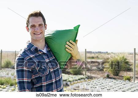 Stock Image - Portrait of young male worker carrying sack on shoulders at plant nursery. Fotosearch