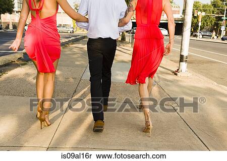 Stock Photography - Rear cropped view of two young women arm in arm with male friend strolling on street. Fotosearch