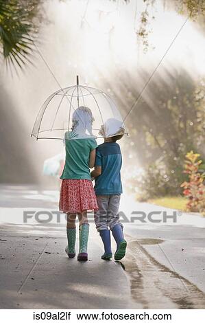 Stock Image - Rear view of boy and girl carrying umbrella on street. Fotosearch
