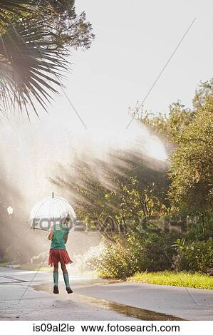 Stock Image - Rear view of girl holding up umbrella and jumping puddles on street. Fotosearch