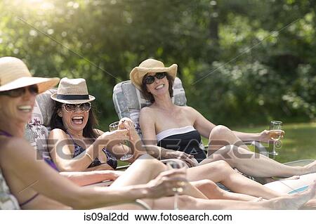 Stock Image - Three mature women relaxing in sun loungers, drinking wine. Fotosearch