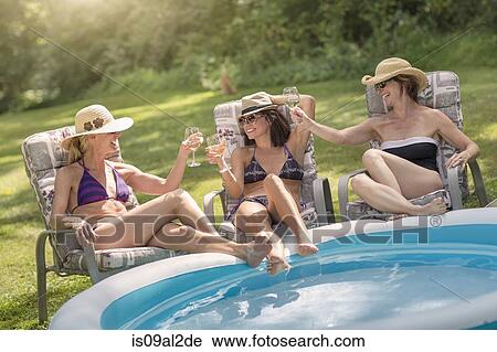 Stock Image - Three mature women relaxing in sun loungers and drinking wine, next to paddling pool. Fotosearch