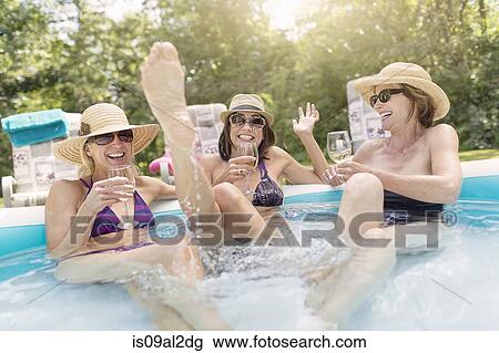 Stock Image - Three mature women sitting in paddling pool, drinking wine. Fotosearch