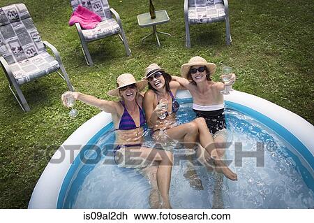 Stock Image - Three mature women sitting in paddling pool, drinking wine, elevated view. Fotosearch