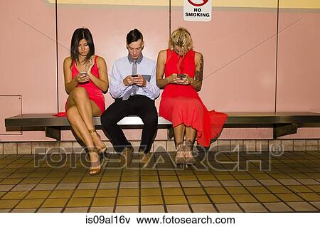 Three young adult friends texting on smartphones whilst sitting in subway station View Large Photo Image Stock Photo - Three young adult friends texting on smartphones whilst sitting in subway station. Fotosearch