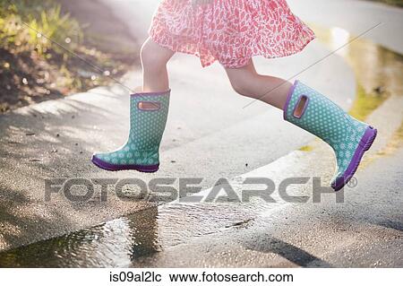 Stock Image - Waist down view of girl jumping puddles on rainy street. Fotosearch